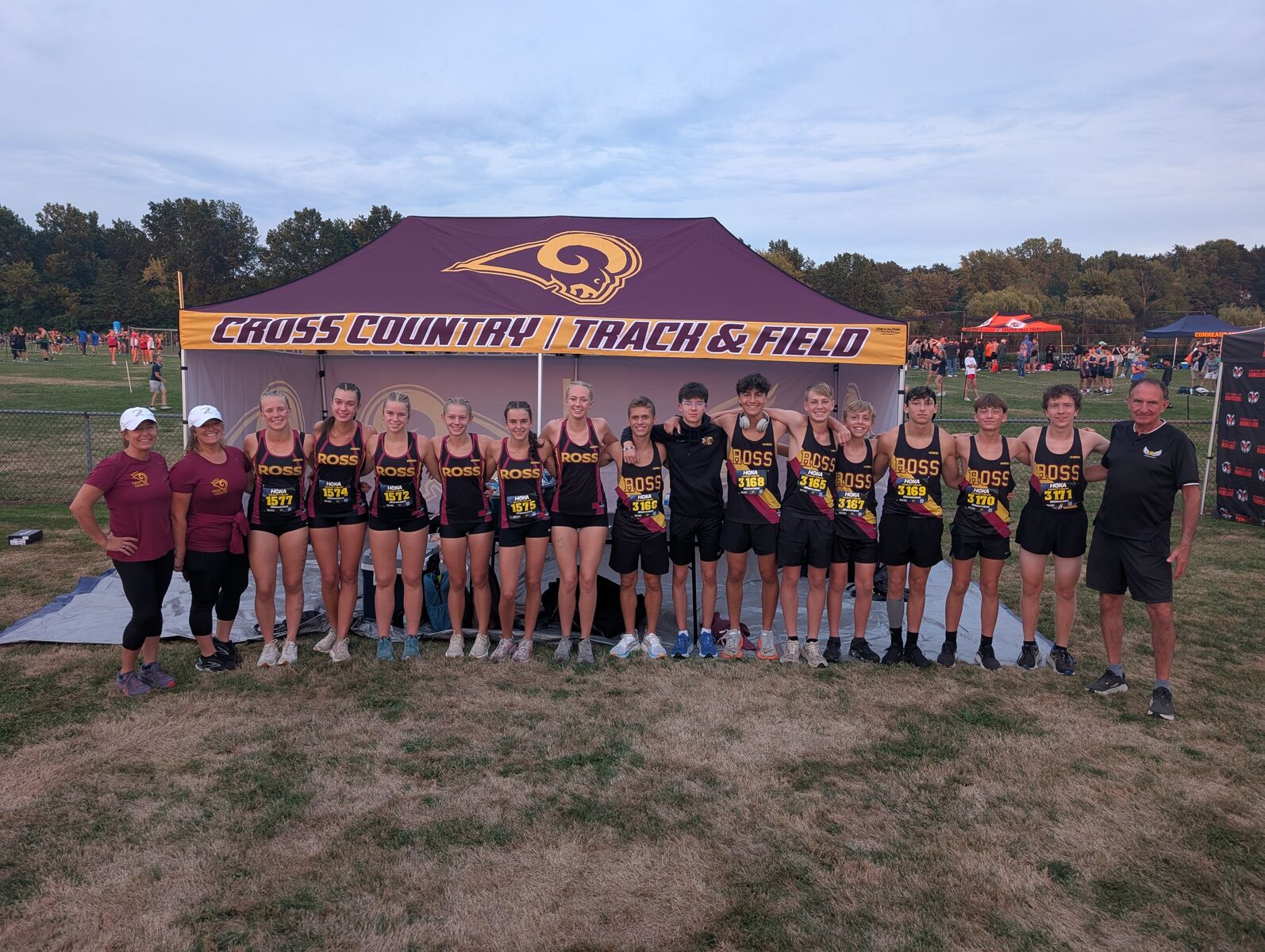 A group of athletes stand in a field in front of a tent that reads Cross County/Track & Field dressed in their cross county uniforms.